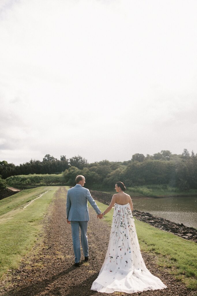 A bride and groom walk hand in hand away from the camera on a dirt path in Hawaii with a small lake on their right side