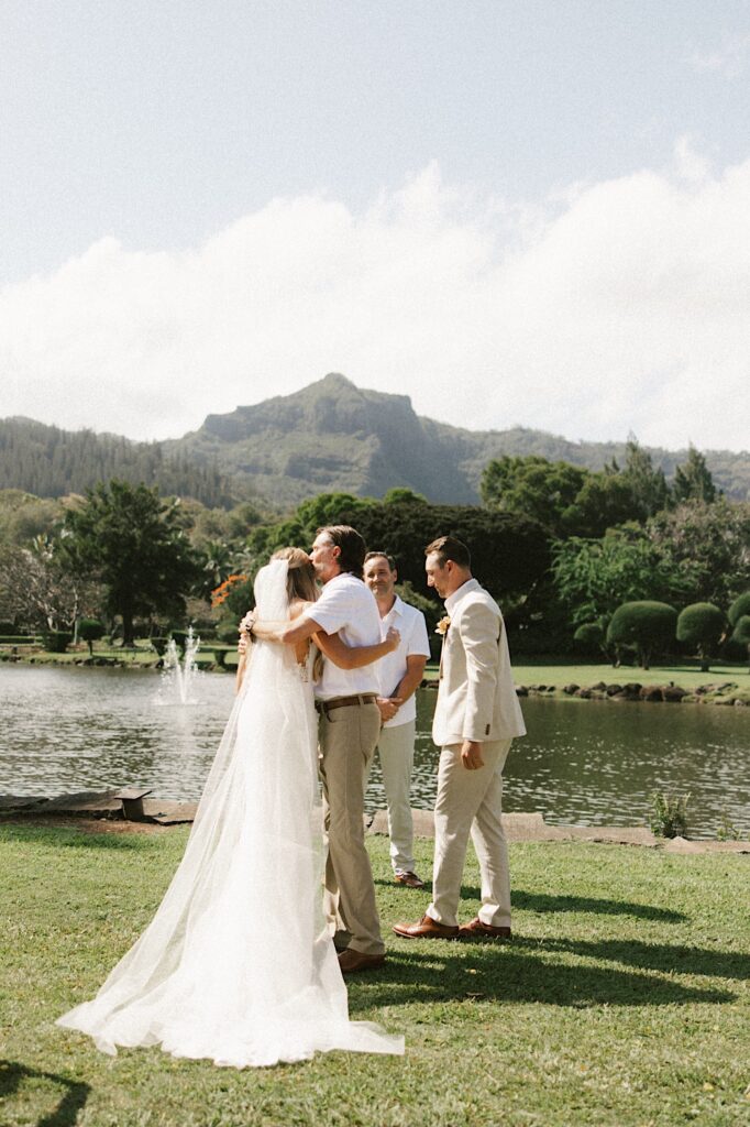 A bride is hugged by a man after he walked her down the aisle of her wedding ceremony with a lake and a mountain behind them, the groom and their officiant stand to the right
