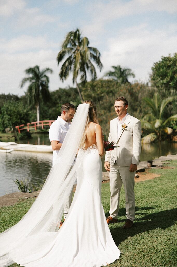 A groom stands looking at his bride who is facing away from the camera during their ceremony in front of a lake in Hawaii