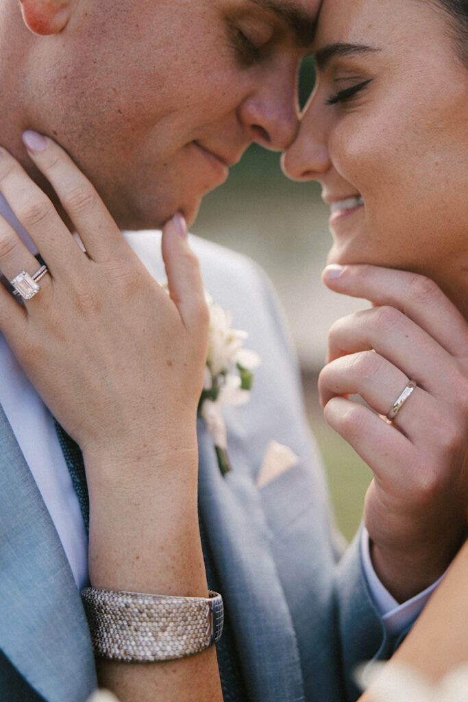 Close up photo of a bride and groom touching their noses together and closing their eyes with their hands with wedding rings on full display touching each others faces