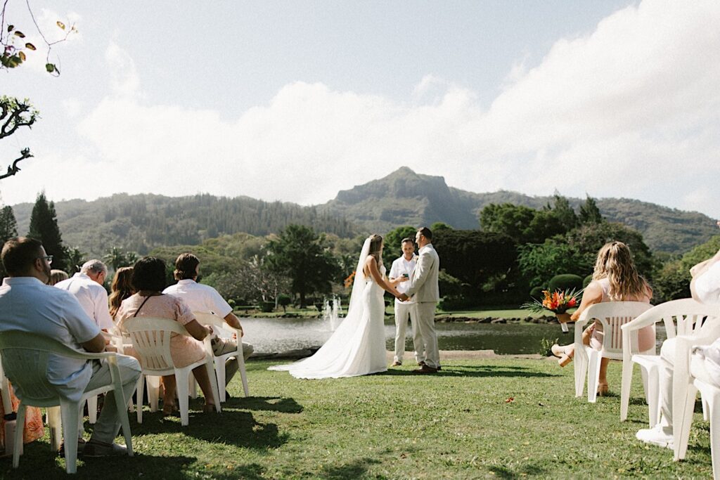 A bride and groom hold hands in front of a lake and mountain during their intimate wedding ceremony on Kauai