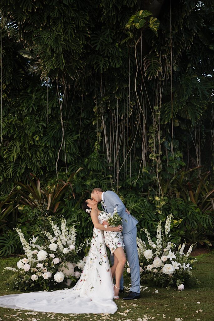 A bride and groom kiss one another while standing next to white flowers and a large tree covered with vines behind them