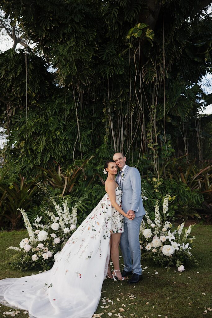 A bride and groom stand together and hold hands while smiling at the camera in front of white floral decor and a large tree covered in vines