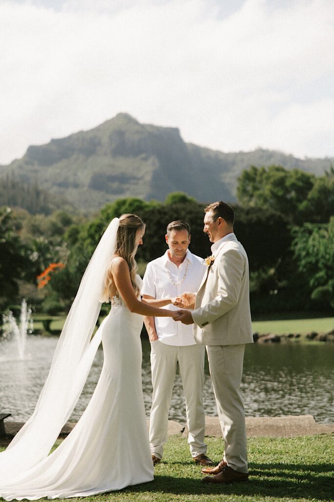 A bride and groom stand holding hands in front of their officiant, a lake, and a mountain in Hawaii