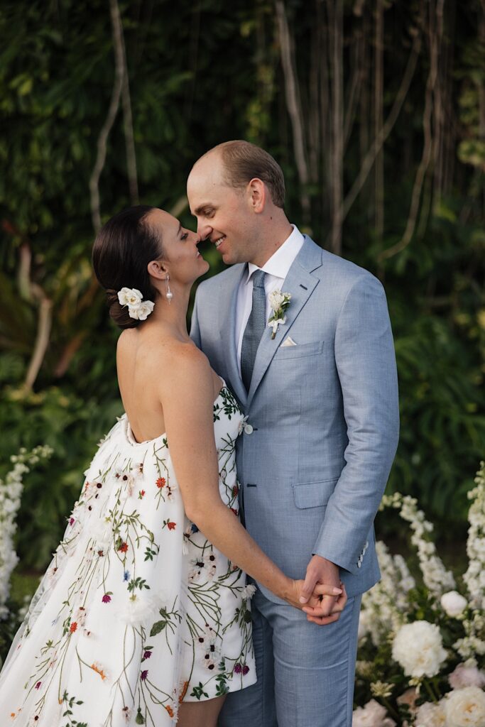 A bride and groom hold hands in front of a tree and white flowers and smile as they are about to kiss