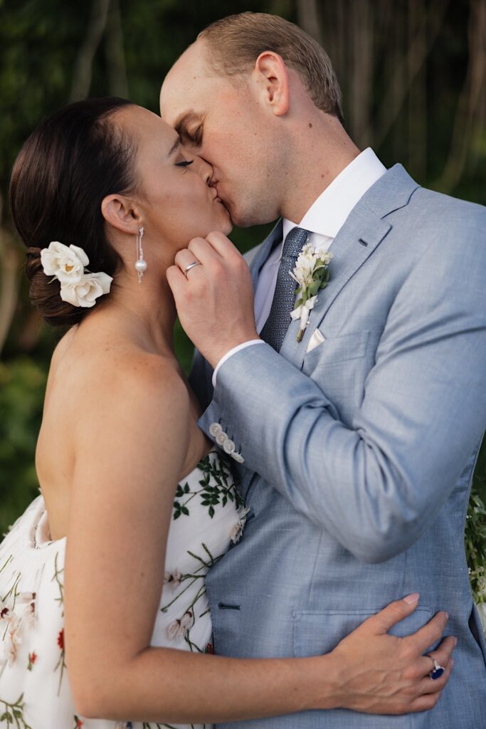 A bride and groom kiss one another and embrace with both the bride's and groom's wedding rings on display