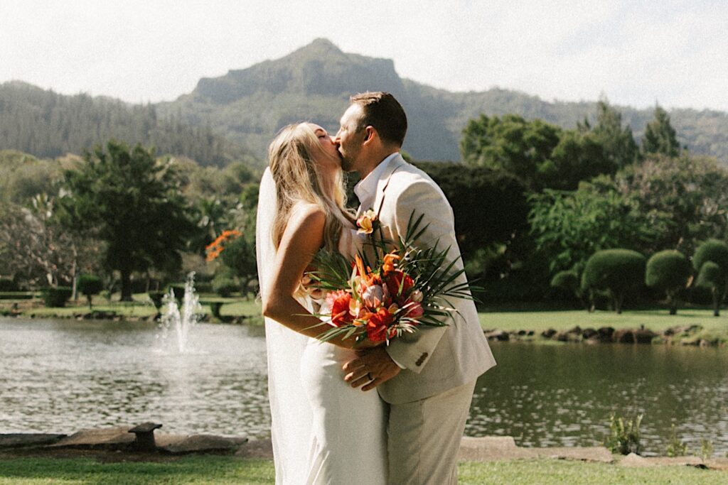 A bride and groom kiss while standing in front of a lake and a mountain on Kauai after their intimate wedding ceremony