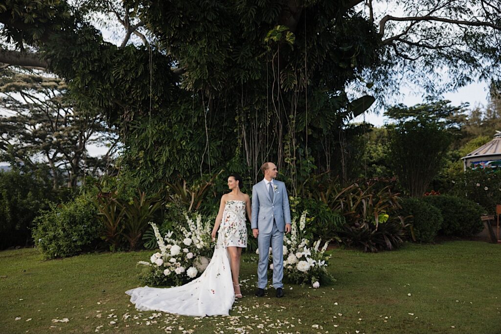 A bride and groom stand next to one another holding hands and looking in opposite directions at their wedding ceremony space at Kukui'ula on Kauai which is covered in white flowers and is in front of a massive tree that is covered in vines