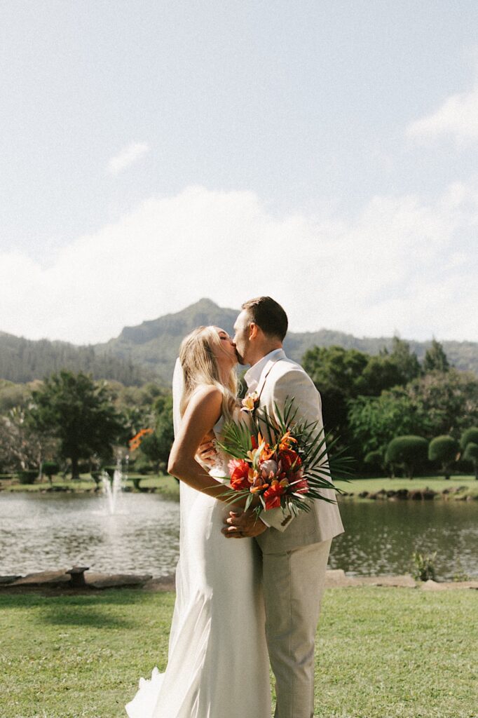 A bride and groom kiss during their wedding ceremony while standing in front of a lake and mountain in Hawaii