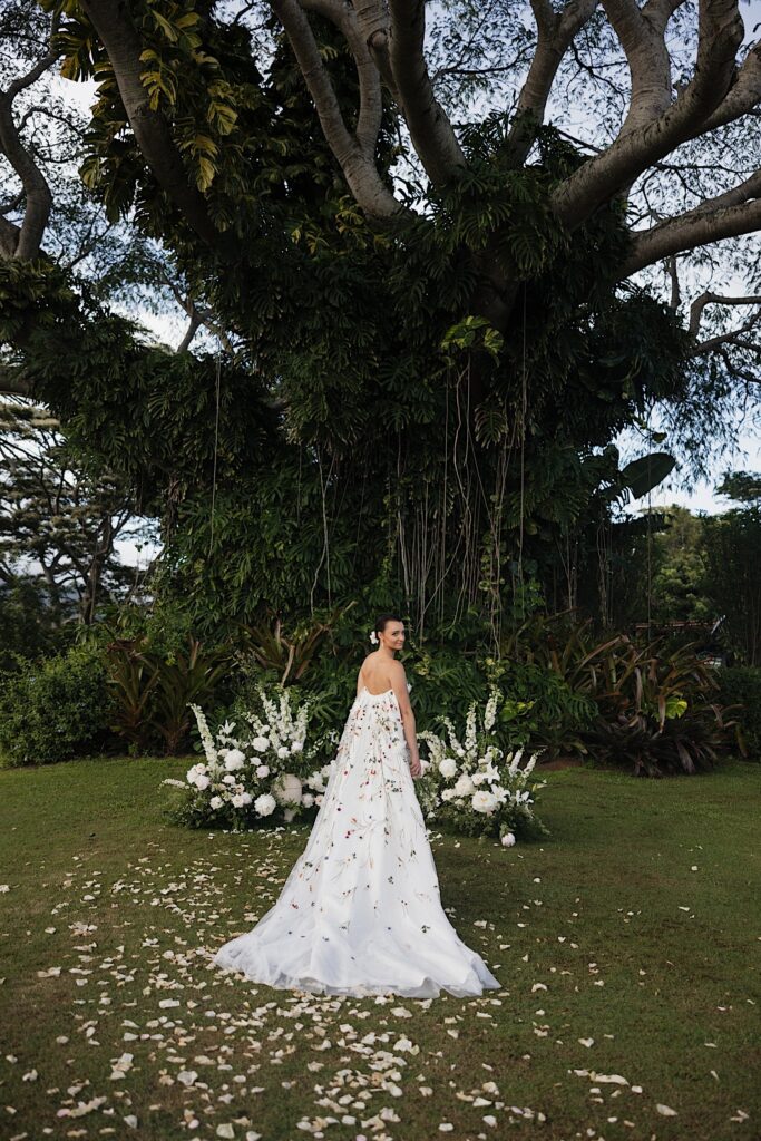 Portrait photo of a bride facing away from the camera and looking over her shoulder at the camera at her ceremony space which is decorated with white flowers and a massive tree behind her