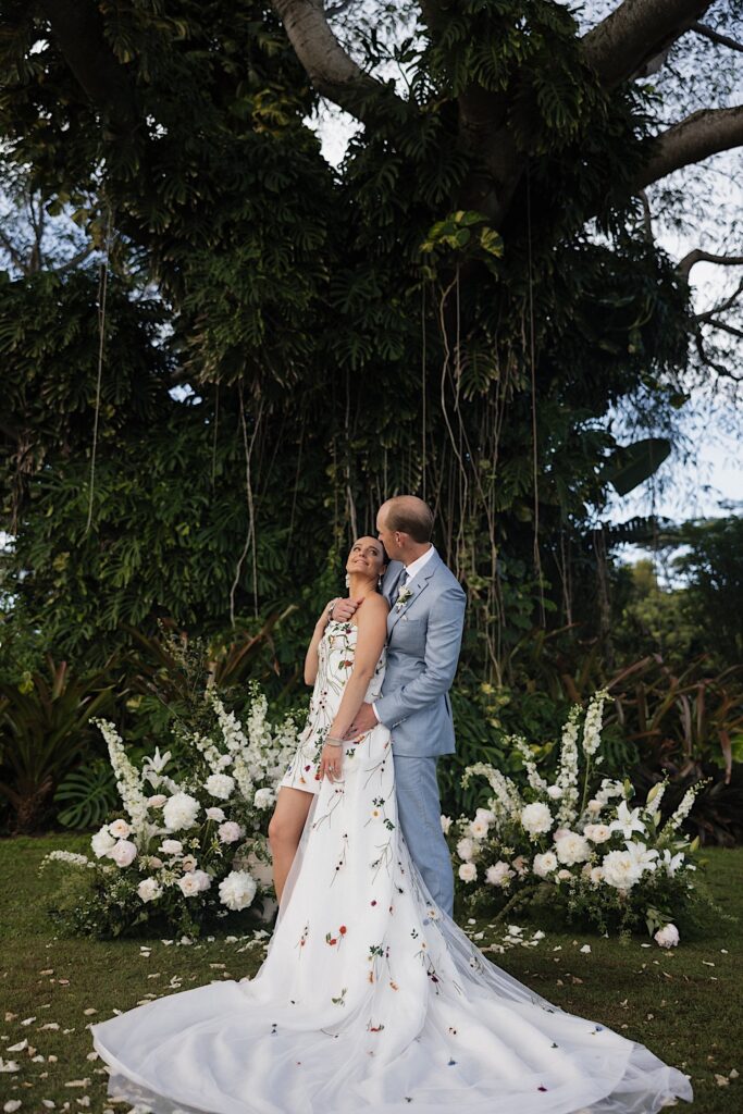 A bride smiles over her shoulder as the groom hugs her from behind as the two stand in front of a large tree covered in vines and white flowers on either side of them