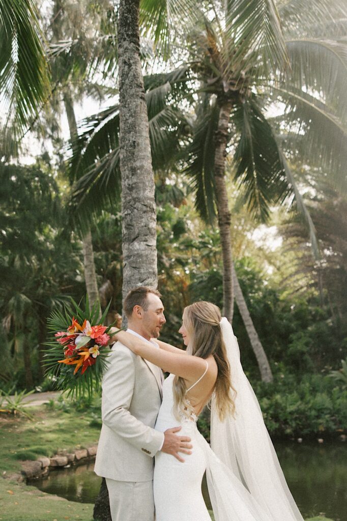 A bride and groom stand facing one another and embrace while standing underneath palm trees