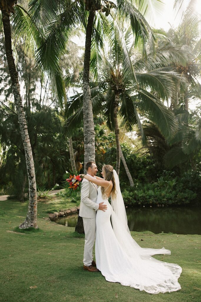 A bride and groom stand facing one another and embrace while standing underneath palm trees