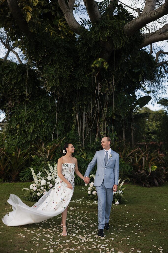 A bride and groom walk hand in hand with one another in front of a massive tree covered in vines towards the camera while looking at one another