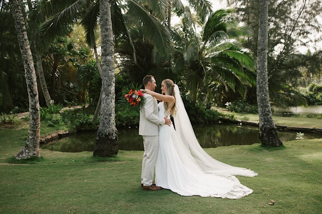 After their intimate wedding ceremony on Kauai a bride and groom stand in front of a small pond and underneath some palm trees as they embrace one another