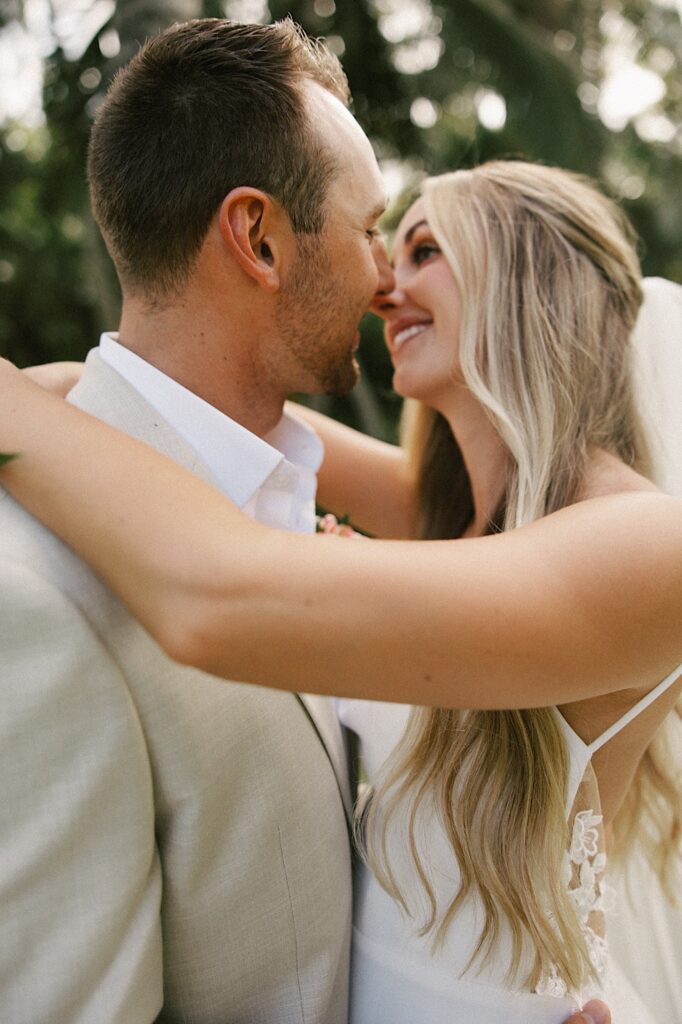 A bride and groom smile at one another as they hug and are about to kiss with palm trees behind them