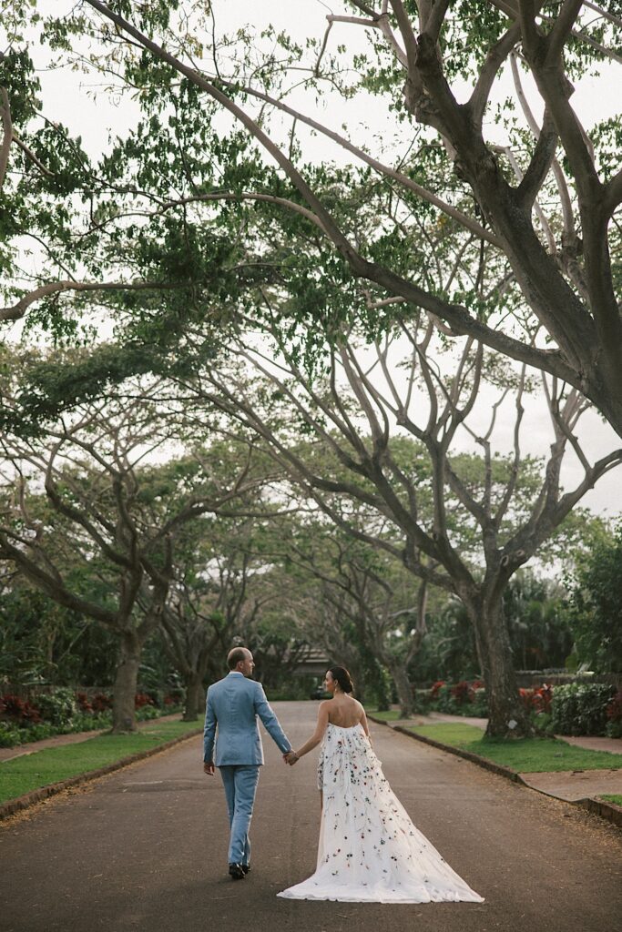 A bride and groom hold hands and walk away from the camera while looking at one another down a road with trees on either side of them