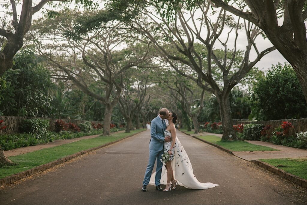 A bride and groom kiss one another on a road with trees on either side of them at their wedding venue Kukui'ula on Kauai