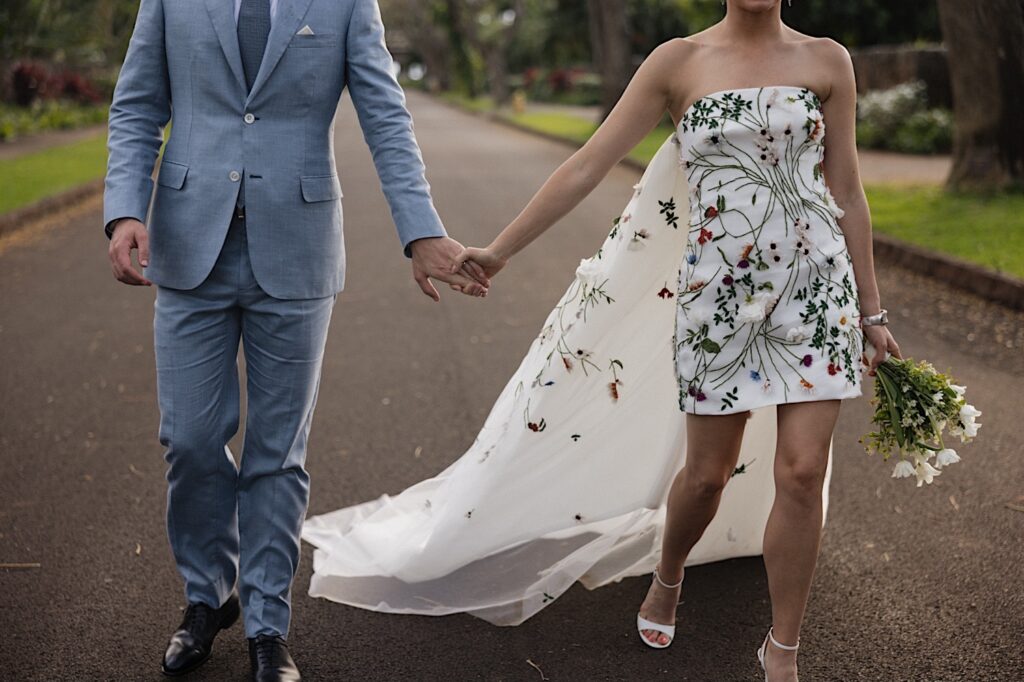 Shoulder down photo of a bride and groom holding hands and walking towards the camera down a street at their wedding venue Kukui'ula on Kauai