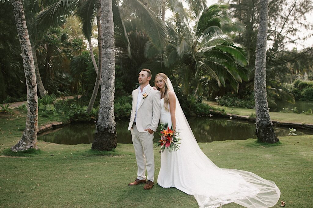 After their intimate wedding ceremony on Kauai a bride and groom stand side by side and pose with a small pond behind them and palm trees above them, the groom is looking to the left while the bride looks at the camera