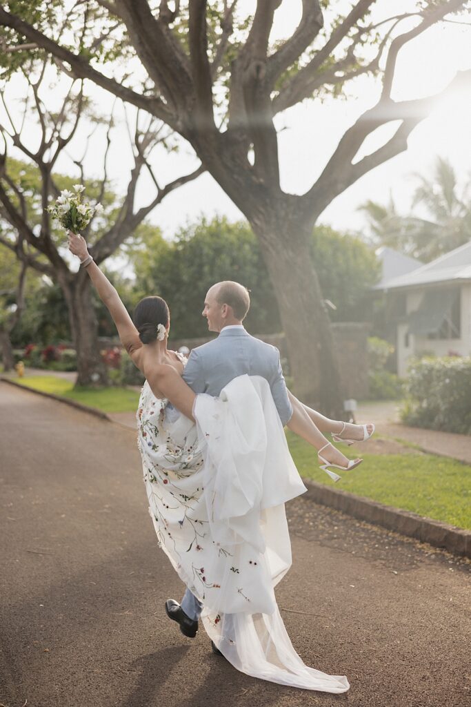 A bride is carried by the groom with her arm and bouquet in the air as the groom walks away from the camera down a street