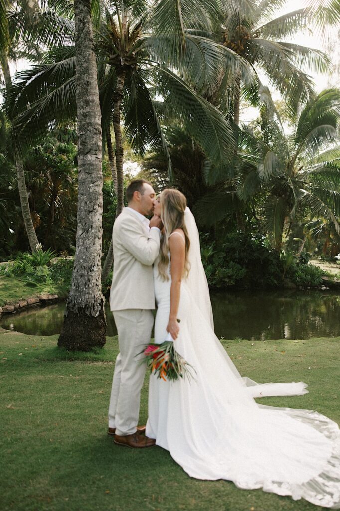 A bride and groom kiss one another while standing in front of a small pond and palm trees