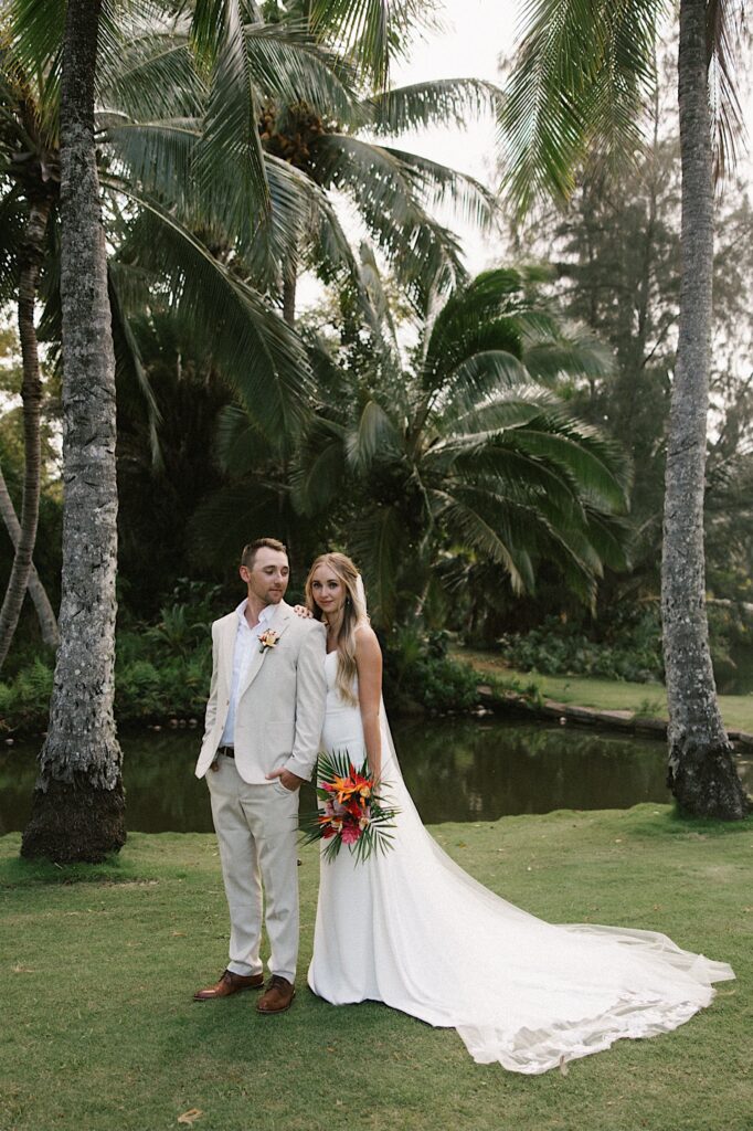 A bride and groom stand next to one another and pose in their wedding attire in front of palm trees and a small pond