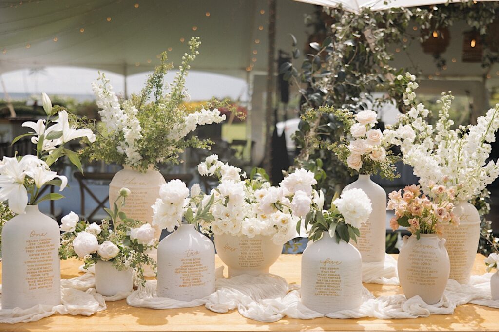 A table set up with white vases with table numbers and guest lists on them, all have white flowers in them