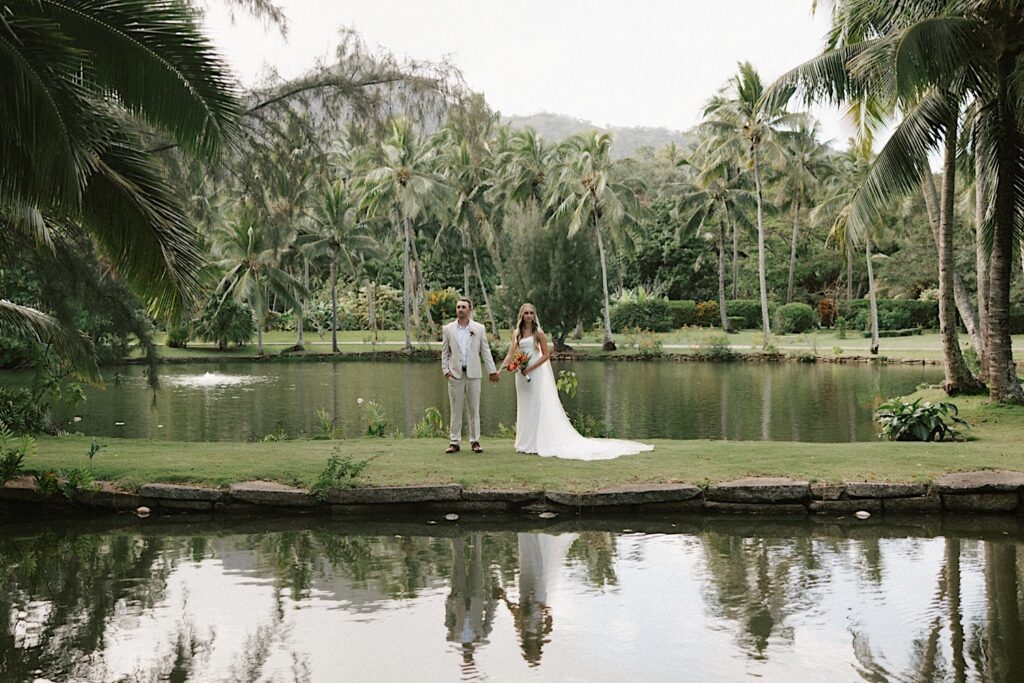 After their intimate wedding ceremony on Kauai a bride and groom stand on a small grass bridge between two bodies of water and hold hands with palm trees behind them