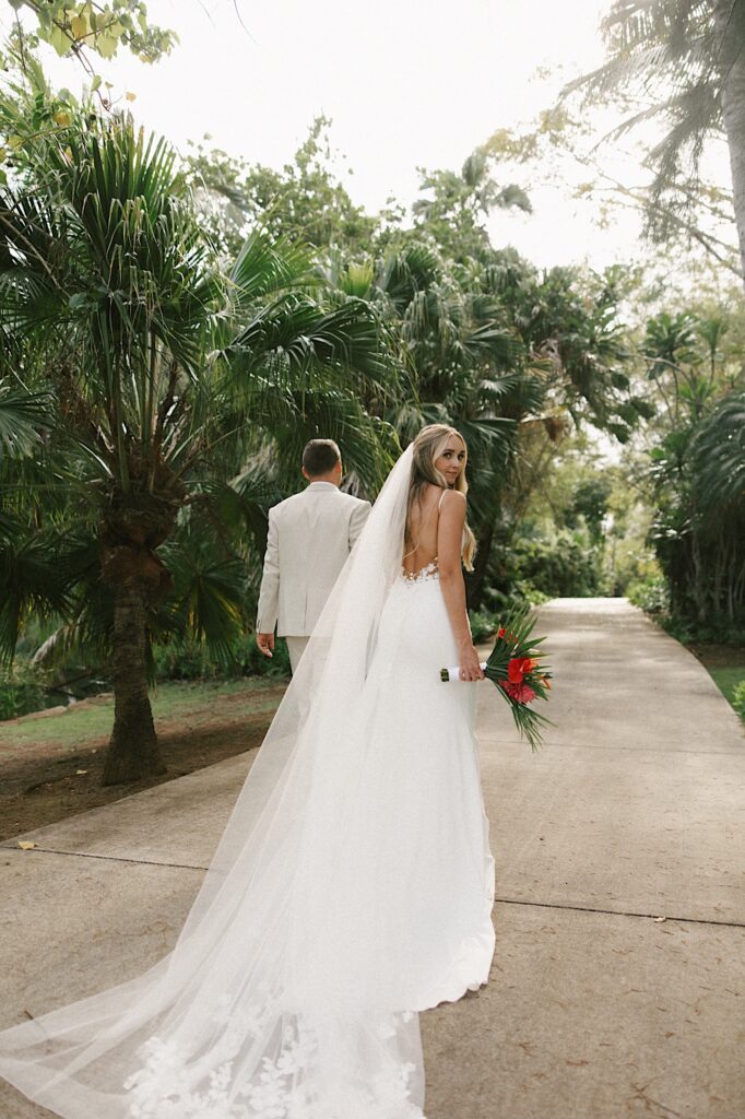 A bride looks over her shoulder behind her back at the camera as her and the groom walk on a path through palm trees