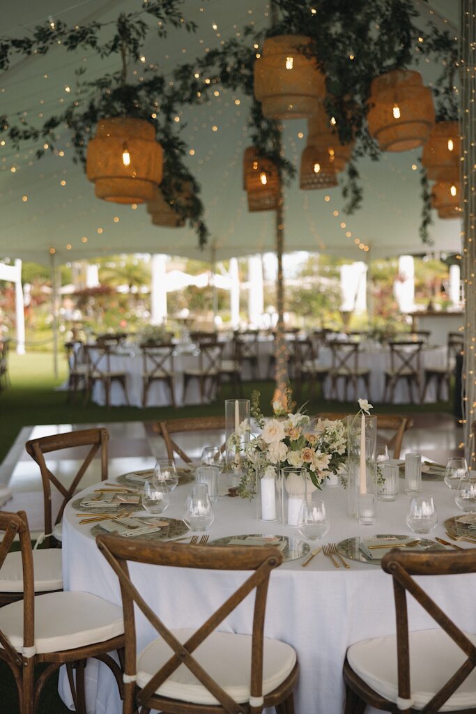 A table sits underneath a tent decorated with greenery and string lights prior to a wedding ceremony