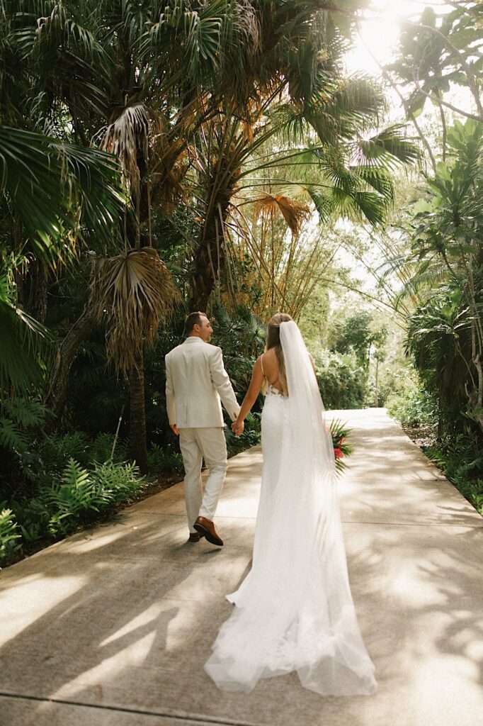 A bride and groom hold hands and walk away from the camera down a path surrounded by palm trees