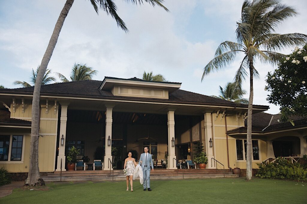 A bride and groom walk hand in hand towards the camera away from a large building towards their wedding reception at Kukui'ula on Kauai