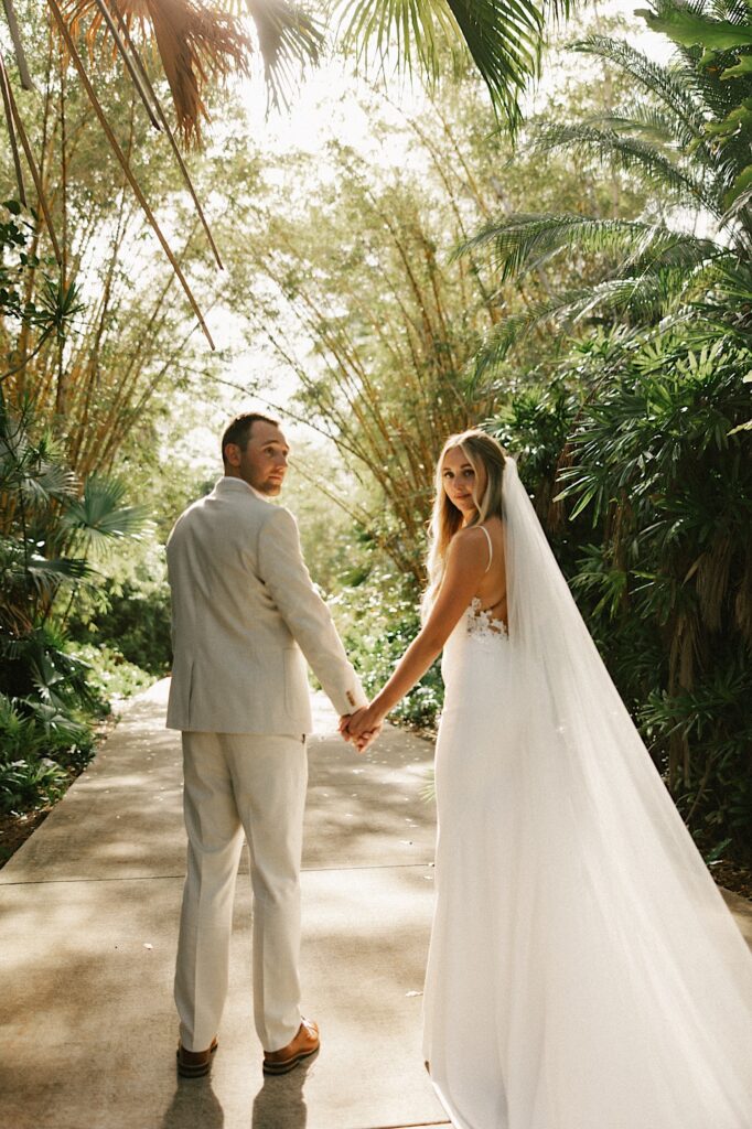 A bride and groom stand on a sidewalk surrounded by palm trees and look back at the camera while holding hands
