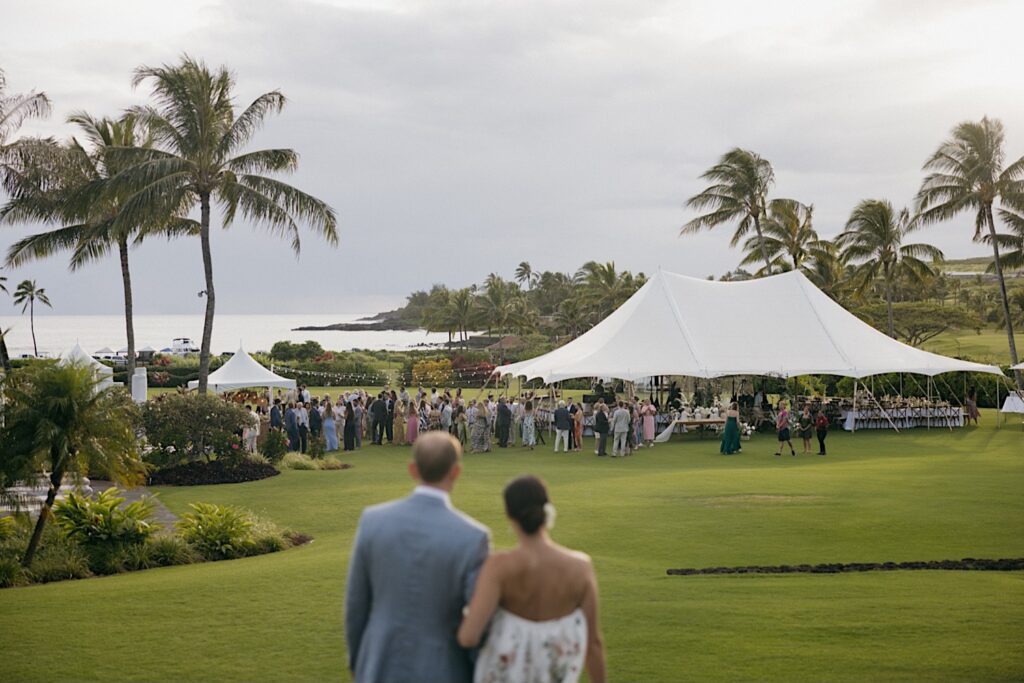 A bride and groom stand next to one another and look out over a large field that has a tent on it and the guests of their wedding reception there waiting for them at their venue Kukui'ula on Kauai