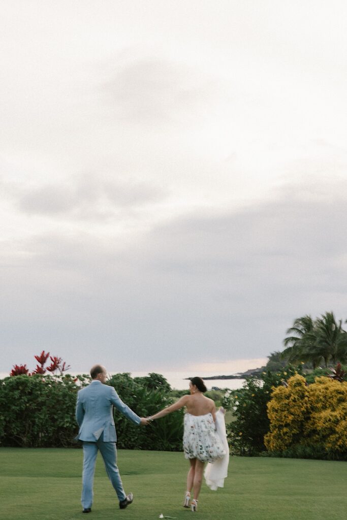 A bride and groom walk hand in hand in an open field towards bushes and the ocean