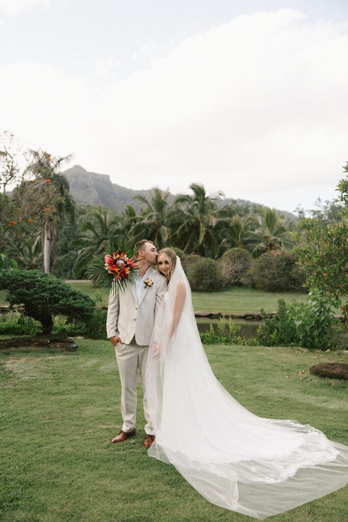 A bride smiles  at the camera while the groom standing next to her kisses her temple, behind them are palm trees and a mountain on Hawaii