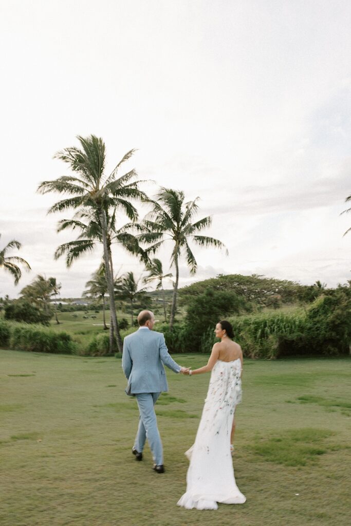 A bride and groom walk hand in hand towards palm trees and the ocean as the two look at one another