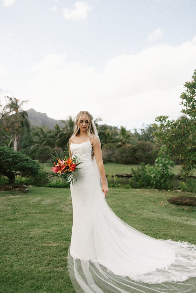 A bride stands in front of a lake and palm trees and looks down at her dress for a portrait photo
