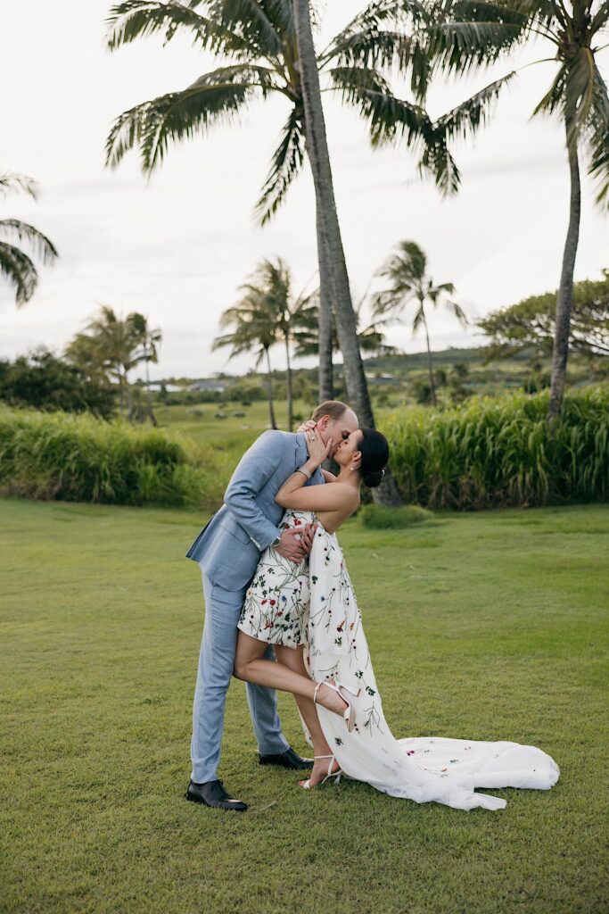 A bride and groom kiss one another with palm trees and the ocean behind them as the bride kicks her leg up behind her