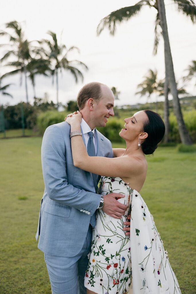A bride and groom embrace and smile at one another with palm trees behind them