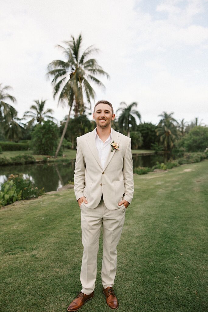 A groom smiles at the camera and puts his hands in his pockets for a portrait photo in front of a river and palm trees