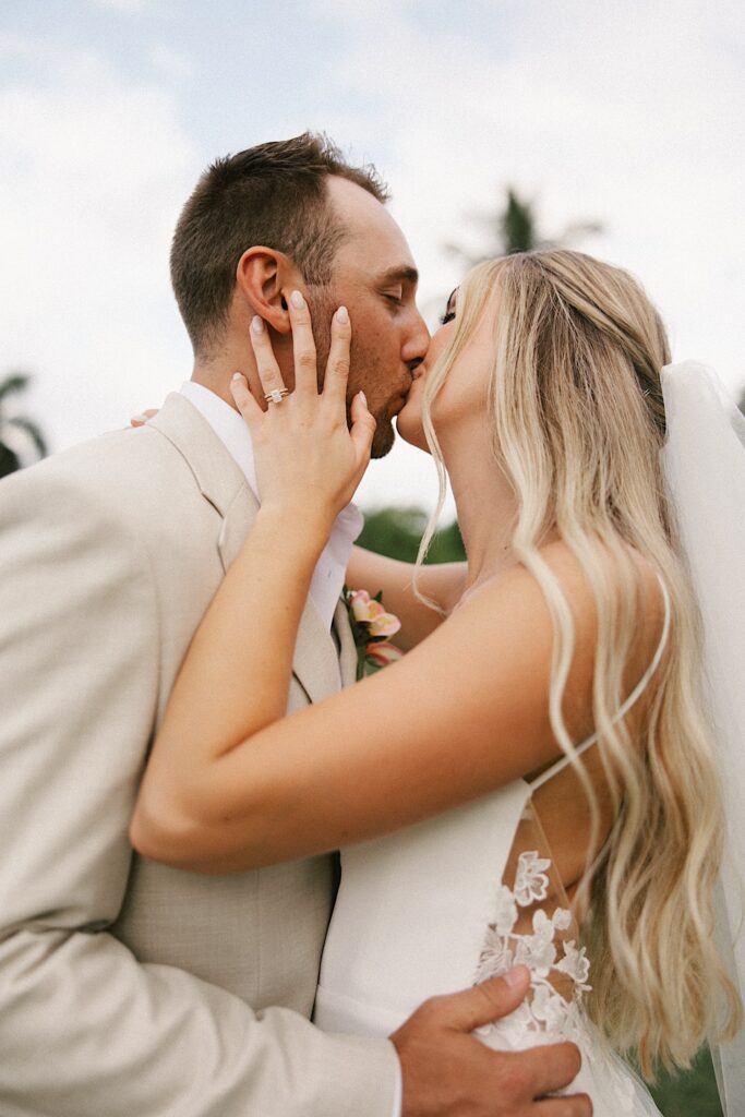 A bride and groom embrace and kiss one another with palm trees behind them