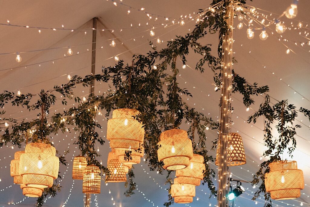 Underneath a tent greenery, string lights, and lanterns are strung to decorate a wedding reception space at Kukui'ula on Kauai