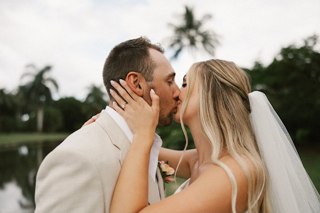 After their intimate wedding on Kauai a bride and groom kiss one another while standing in front of a river and palm trees