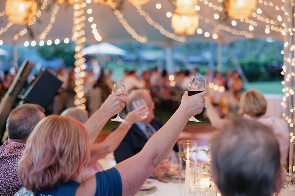 Guests of a wedding underneath a tent decorated with string lights raise their glasses for a toast