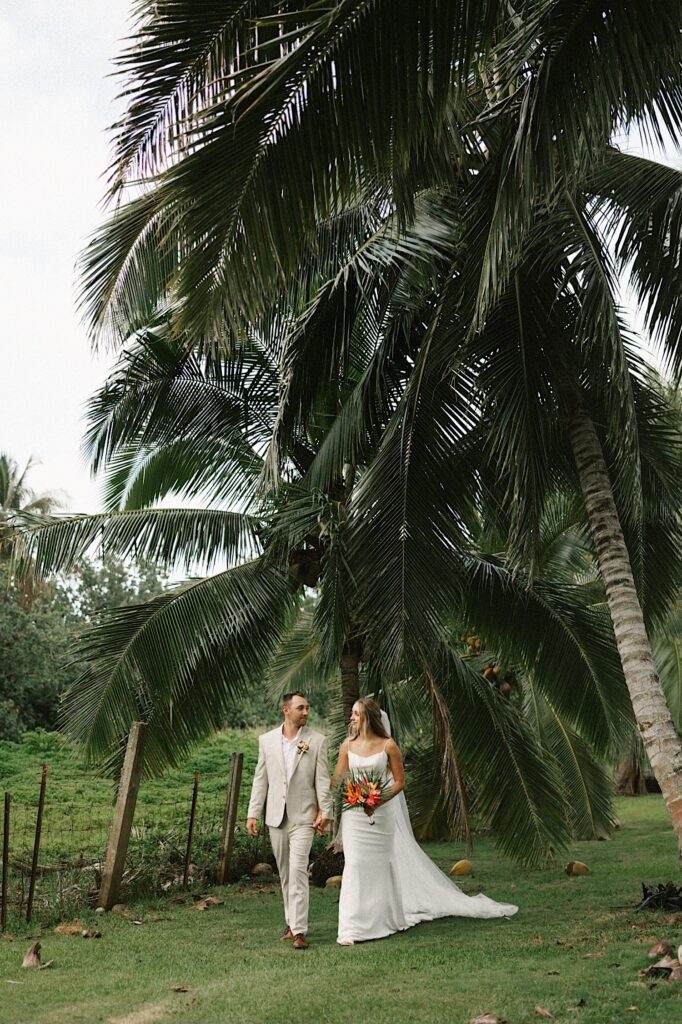 A bride and groom walk hand in hand towards the camera and look at one another in front of a palm tree