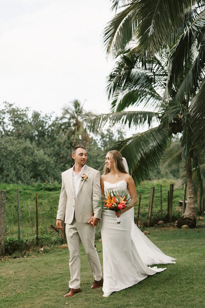 A bride and groom hold hands and look at one another while walking towards the camera in front of palm trees