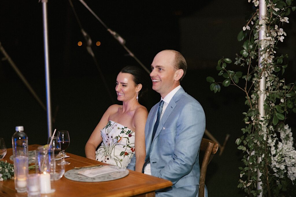 A bride and groom sit next to one another at a head table and smile during a speech at their wedding reception at their venue Kukui'ula on Kauai