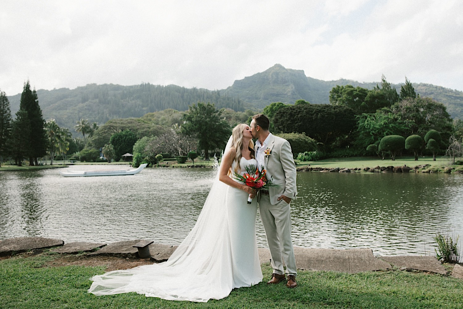 A bride and groom kiss one another in front of a lake, palm trees, and a mountain after their intimate wedding on Kauai